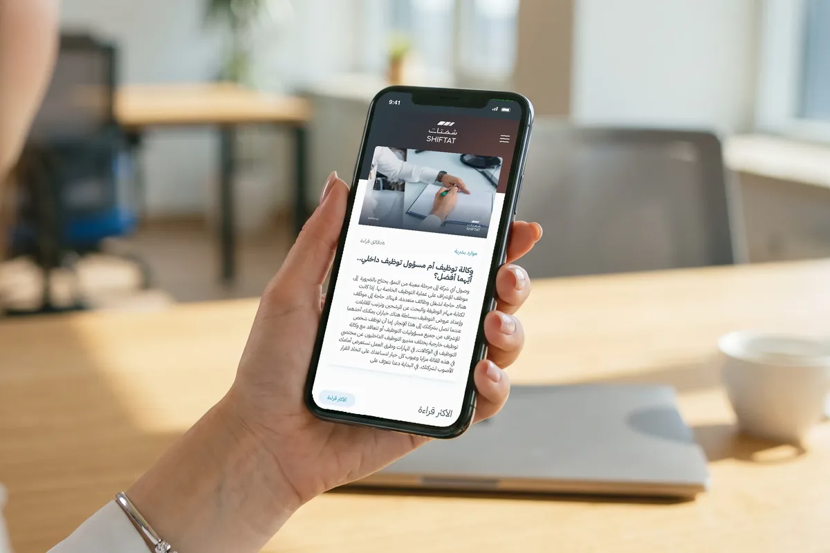 A hand holding a phone with the same Shiftat blog article open, in Arabic